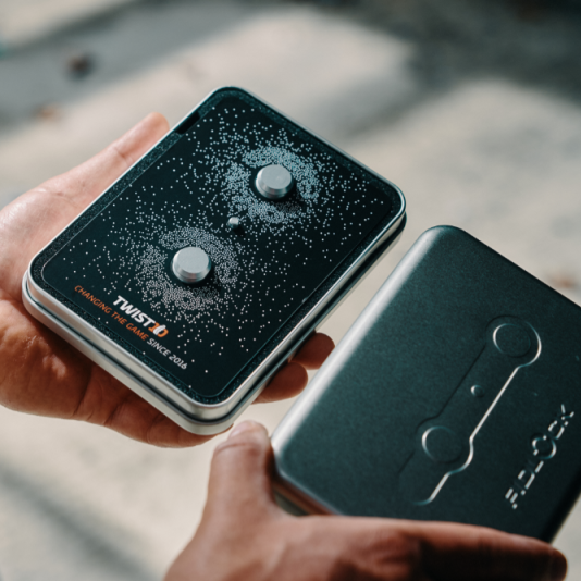 A person holds two black rectangular electronic devices outdoors. The device in the hand has a textured black face with two round silver buttons and visible branding that reads TWISTO.