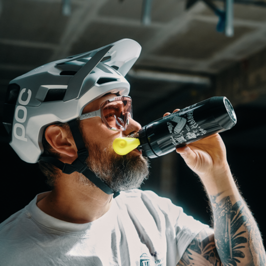 A man wearing a white POC mountain bike helmet and safety glasses drinks from a black can of energy drink with a yellow straw in a dark indoor setting.