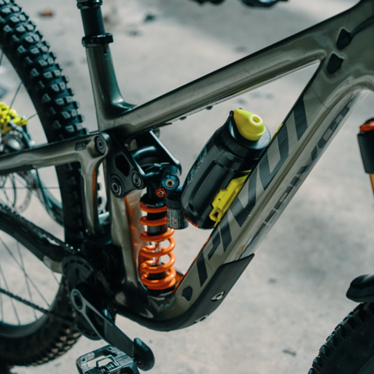Close-up of a mountain bike frame and suspension components, showing a matte dark gray frame with orange rear shock, gold front fork, and a black-and-yellow water bottle mounted on the frame.