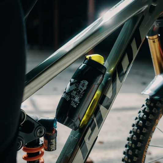 A close-up shot of a black and yellow mountain bike water bottle mounted on the frame of a full-suspension dirt bike, with an orange front fork and knobby tire visible in a blurred outdoor setting.